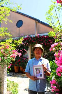 妻・由美子さんの写真を手に庭に立つ居場英則さん